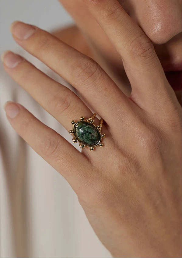 Hand wearing a gold ring with a green stone on a neutral background