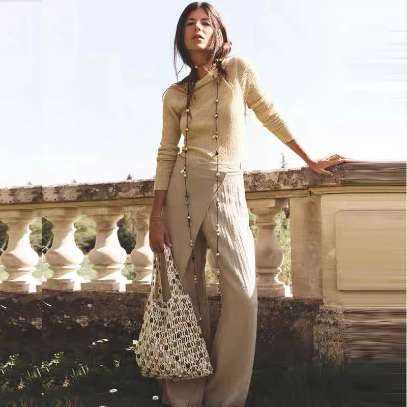 Woman in beige outfit holding a patterned bag, standing against a stone wall with greenery.