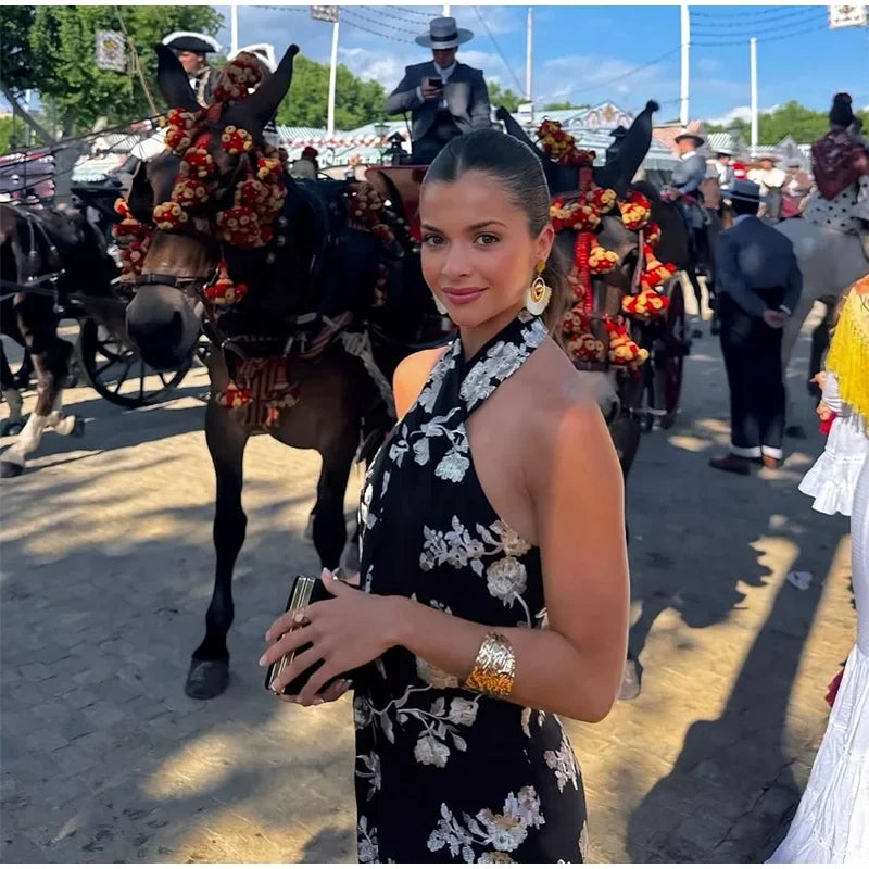Woman in a floral dress standing in front of decorated horses with people in the background.