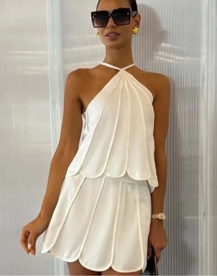 Woman wearing a white halter neck dress with sunglasses and gold jewelry against a neutral background